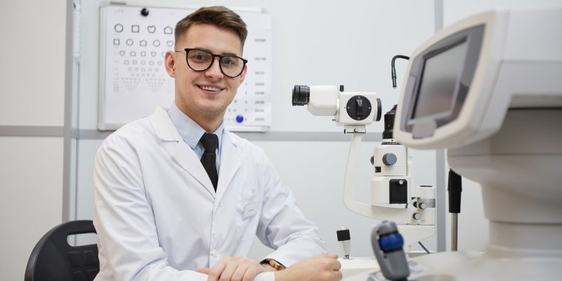 portrait-male-optometrist-smiling-camera-while-posing-workplace-by-optic-equipment (1)-min